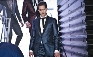 A male model in a sparkly navy suit coming down some stairs, people in suits following