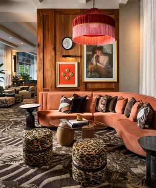 hotel lounge lobby area with wooden panelled wall, a large pink fringe light hung above an orange velvet curved couch with little leopard print ottoman stools and patterned carpet