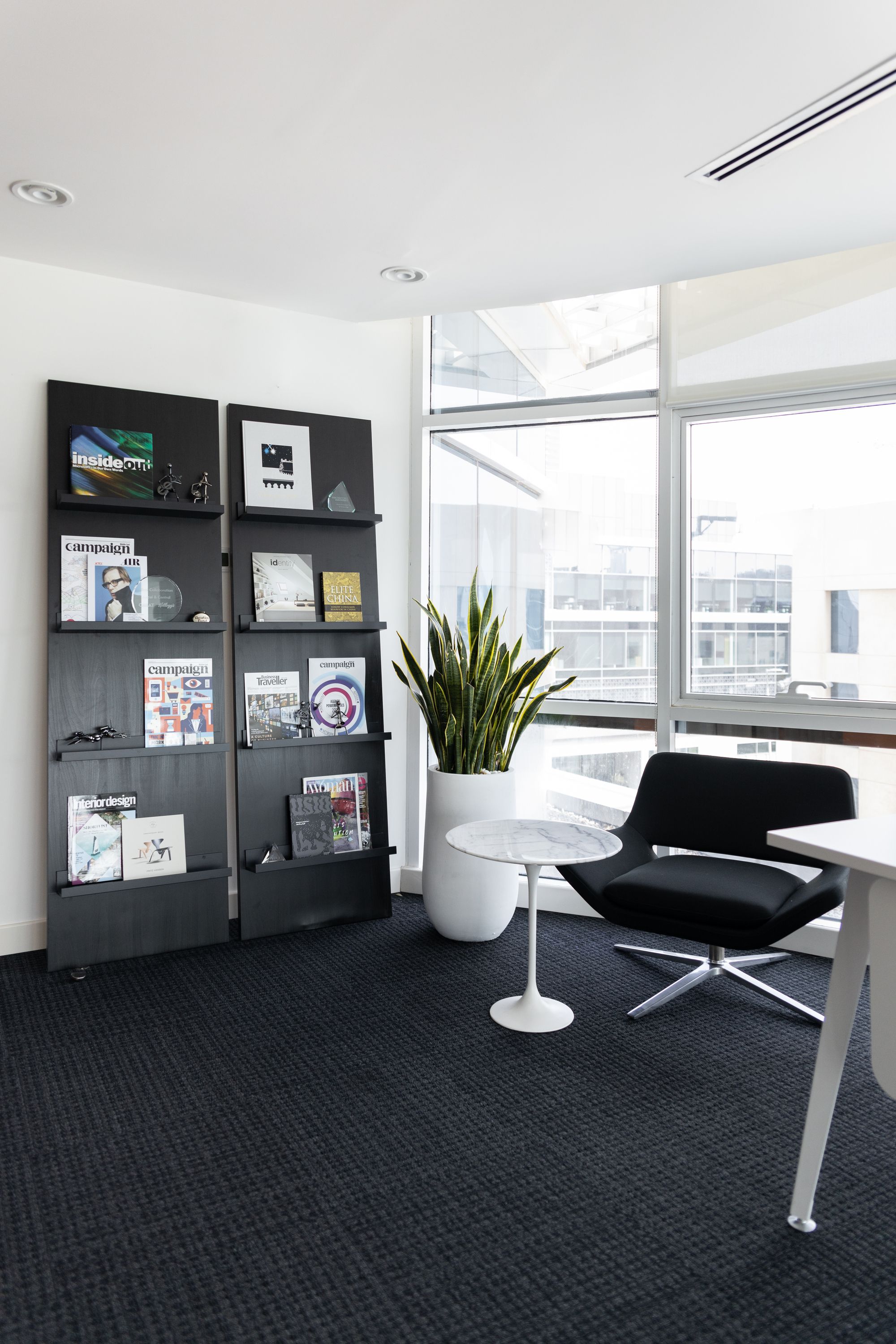 A white office room with black carpets and two large black magazine shelves leaning against the wall. There is also a white vase with a large green plant in it, a white marble table, and a black office chair.