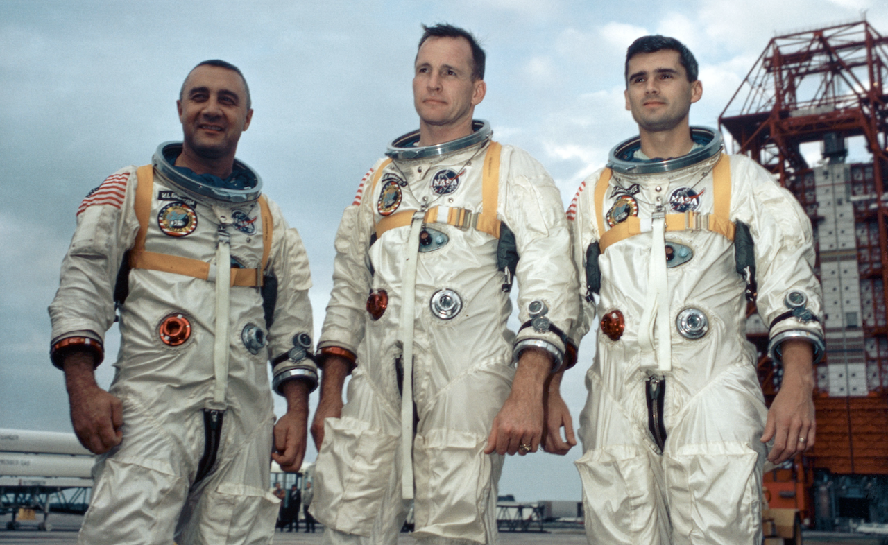Three astronauts standing in spacesuits with a launch tower behind