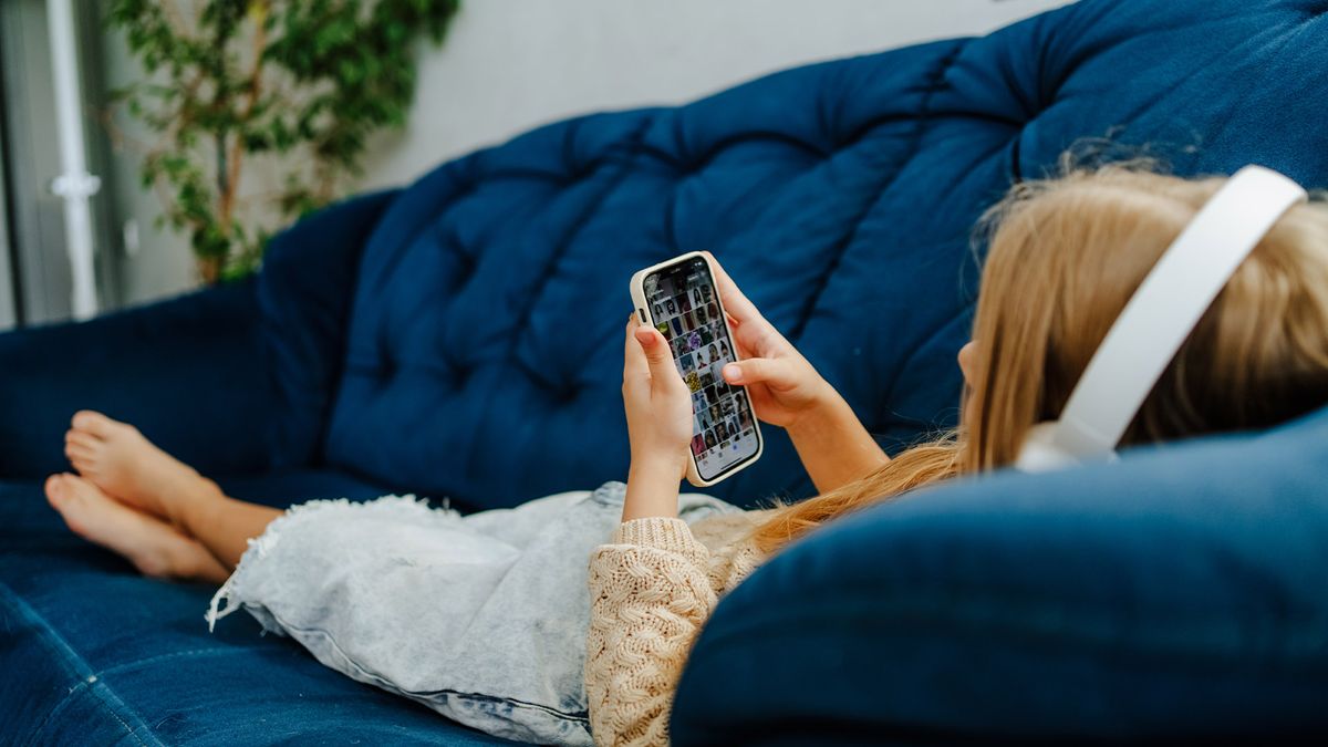 Rearview of a barefoot girl in headphones playing games and listen to music, lying on a sofa indoors.