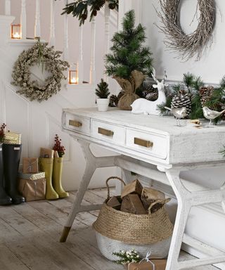 White painted hallway table with pine cones and pine tree foliage on top and woven basket with logs underneath
