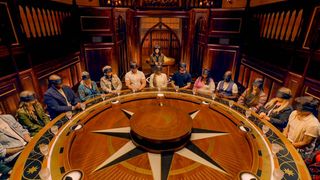 a group of blindfolded people sit around a round table with a star motif, as a woman (claudia winkleman) stands behind them, in 'the traitors u.k.'