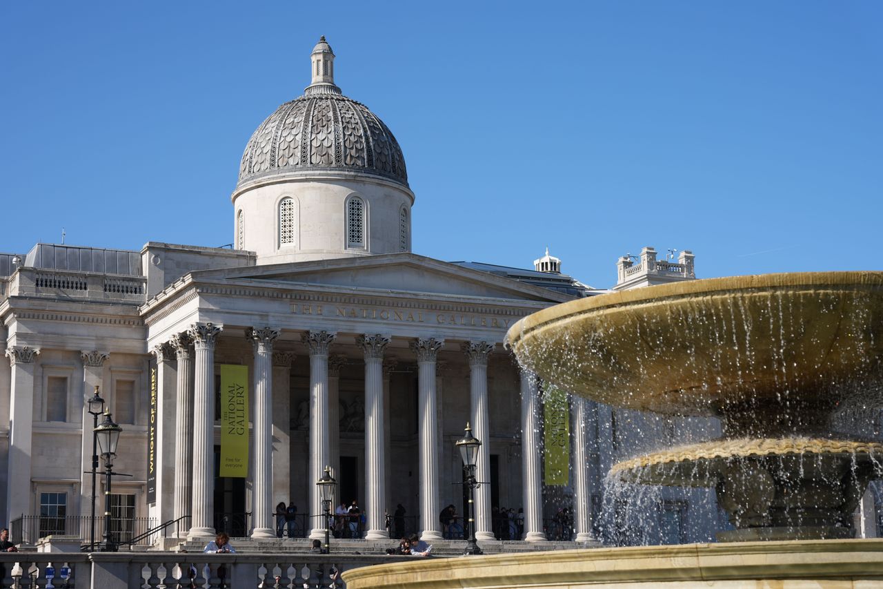 The National Gallery and Trafalgar Square fountain under a clear blue sky.
