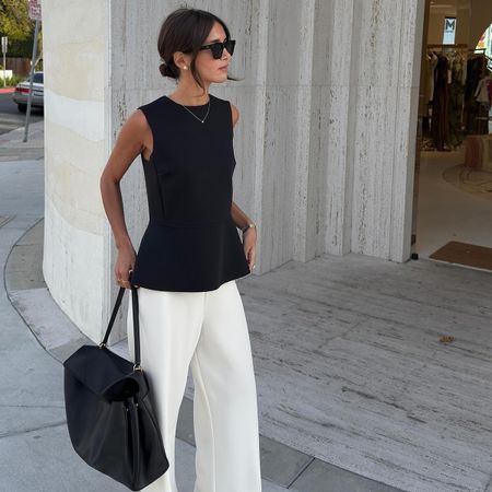 A woman wears a black top, white pants, and a black top-handle bag.