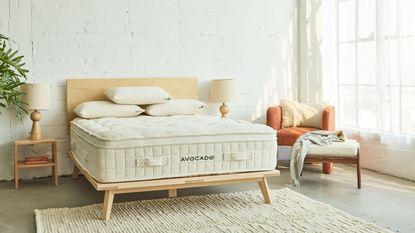 Avocado Luxury Organic Mattress on a light wood bedframe in a white bedroom with wood flooring, exposed brick walls painted white, and large windows to the right.