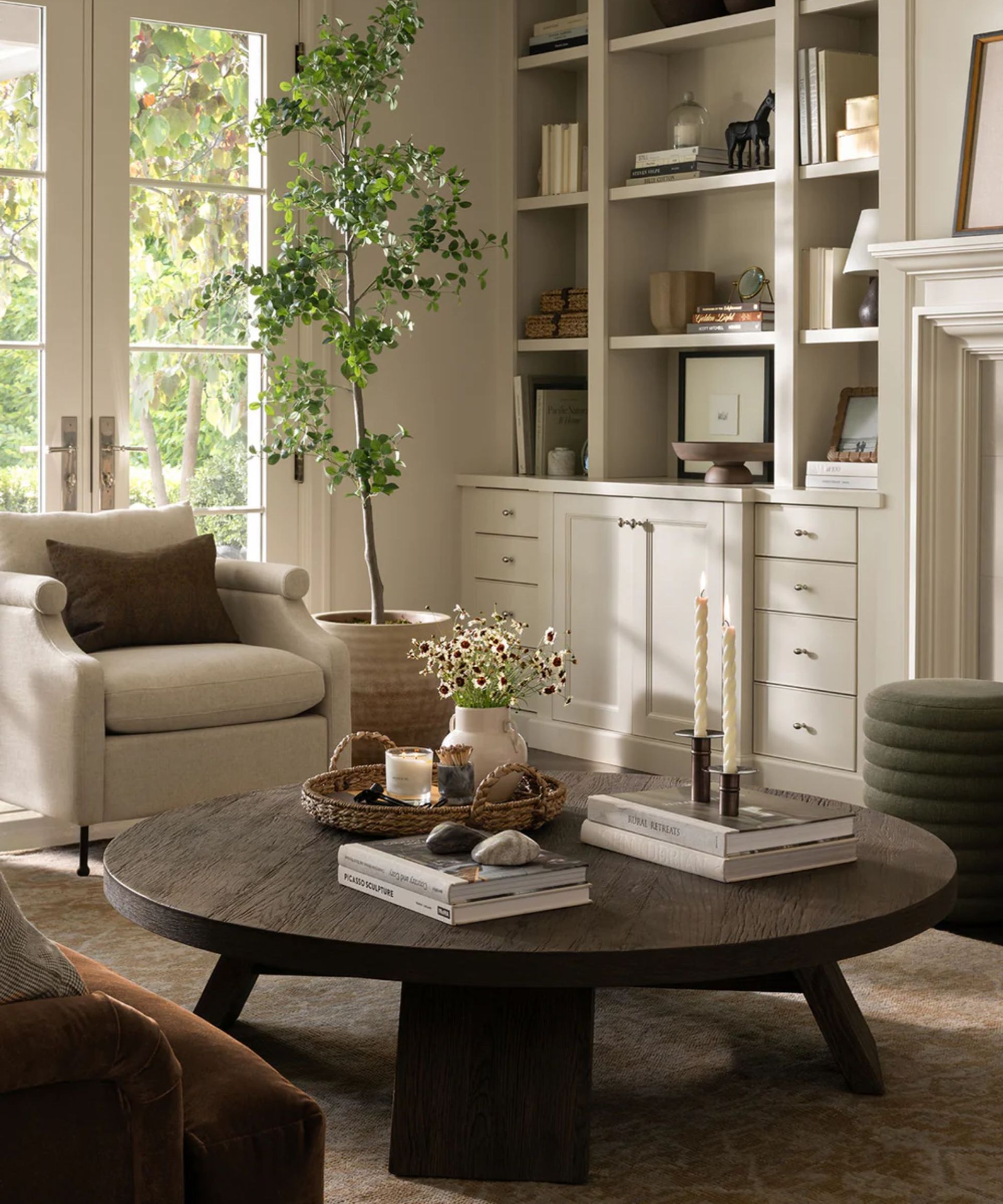 neutral farmhouse living room with a dark wood round coffee table styed for spring with a woven tray scented candles, books and a vase with daisies
