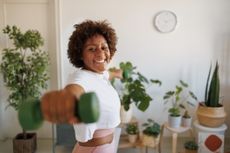 woman facing the camera punching forward with one arm and holding a green lightweight dumbbell. lots of indoor plants are in the background. 