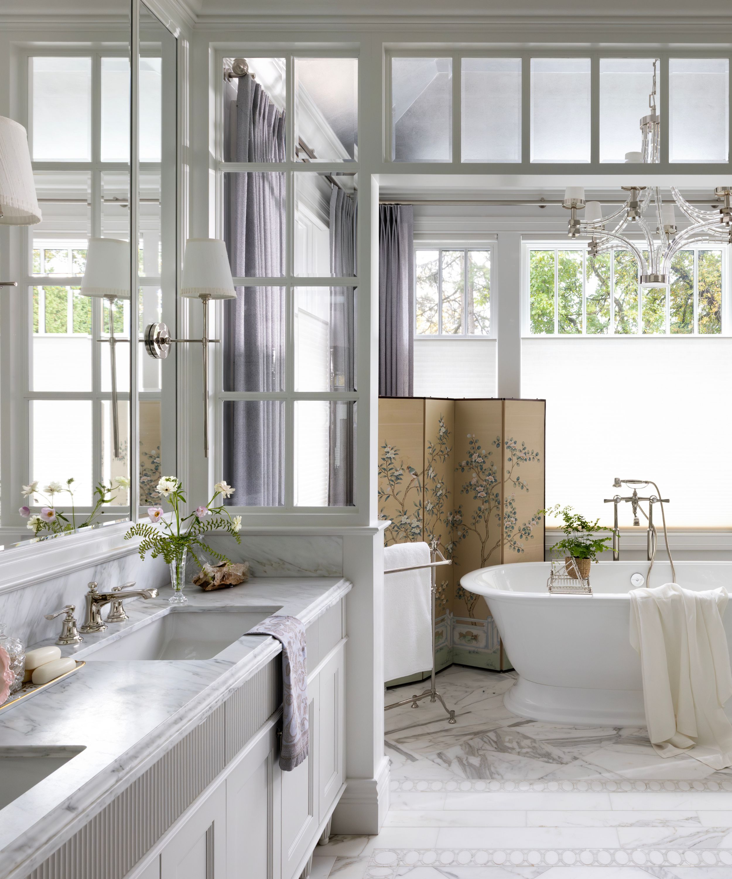 Bright luxury bathroom with a white freestanding tub, marble floors, lavender curtains, and a decorative folding screen under a glass-paned ceiling.