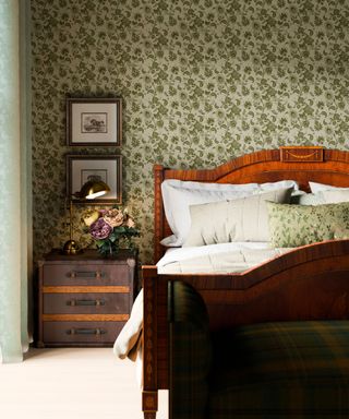 A bedroom with a green floral wallpaper, a bedside table with flowers on it and a dark brown wooden bed with white sheets on it