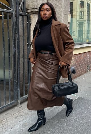 A cute work winter outfit is shown in a photo of a woman standing on the sidewalk in Oslo wearing a black turtleneck sweater under a brown suede blazer with a brown leather skirt, black knee-high boots, and a black handbag