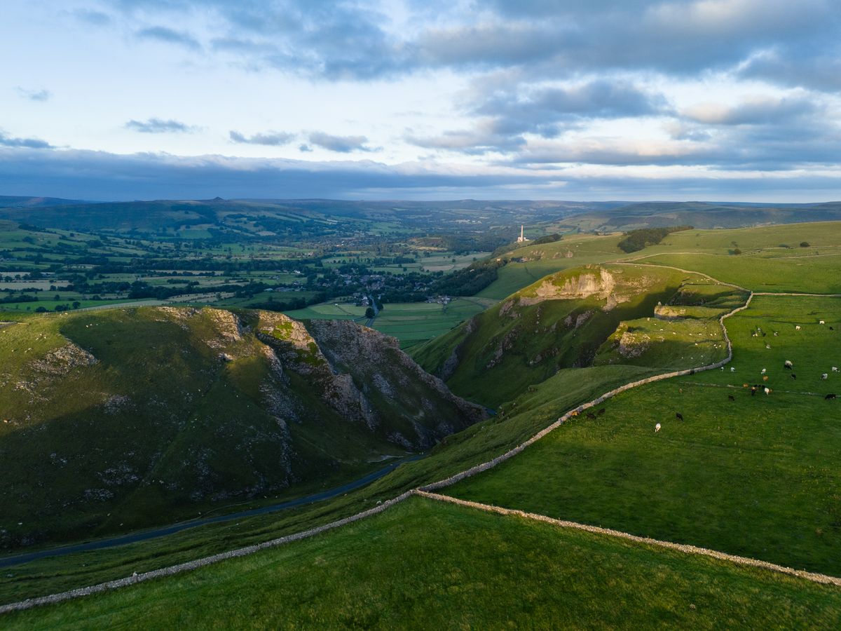 Image taken by DJI Air 3 (a camera drone). Here the focus is on a road winding through the green valleys. In the distance are rolling green hills dotted with dark green trees and a gray, cloudy sky. Just to the right of the image you can see some cows in a field.