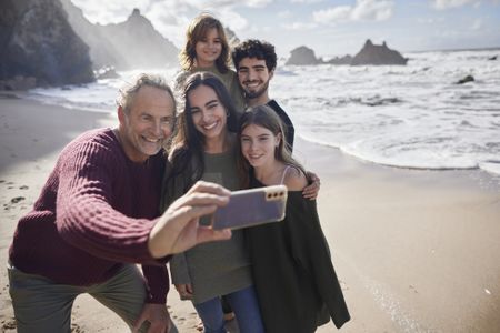 An older man takes a selfie of his family on the beach. 