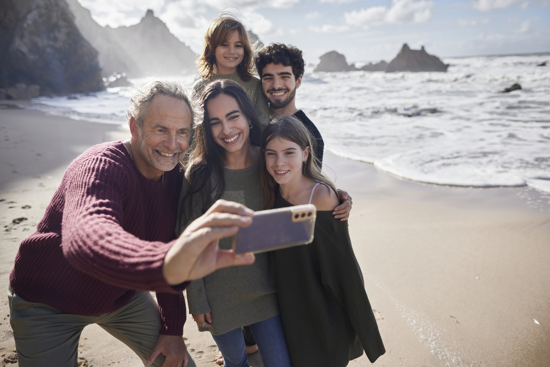 An older man takes a selfie of his family on the beach. 