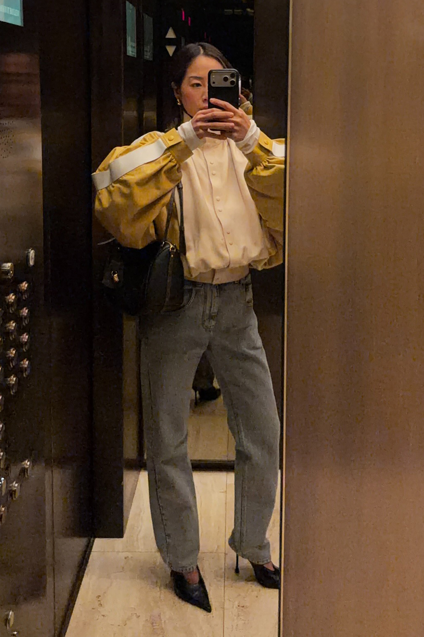 a woman wearing a butter yellow two-tone bomber jacket, blue jeans, and black heels