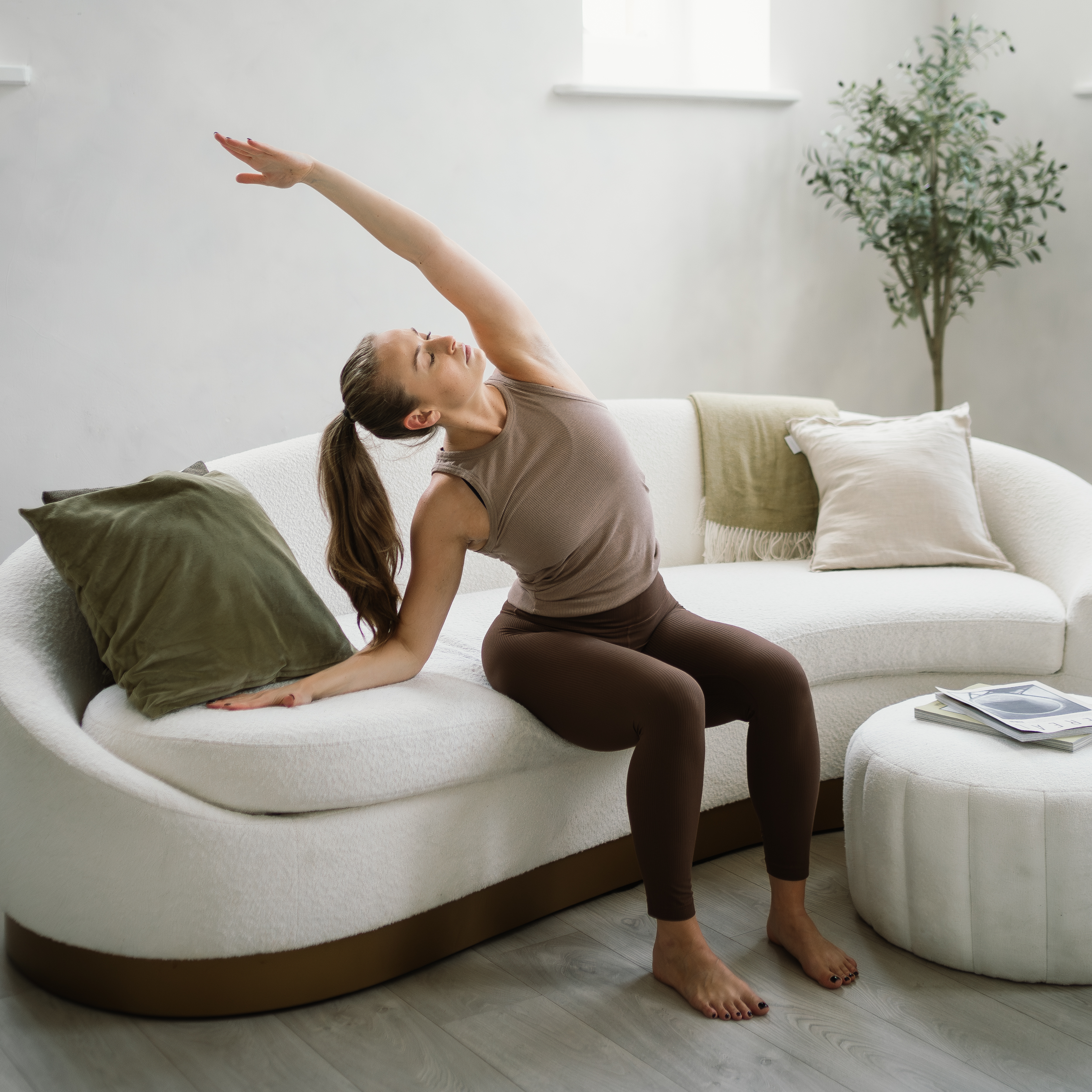 A woman sits on the edge of a curved couch performing a seated side stretch. Her knees are bent, feet flat on the floor as she reaches up and over with her left hand, left arm straight.