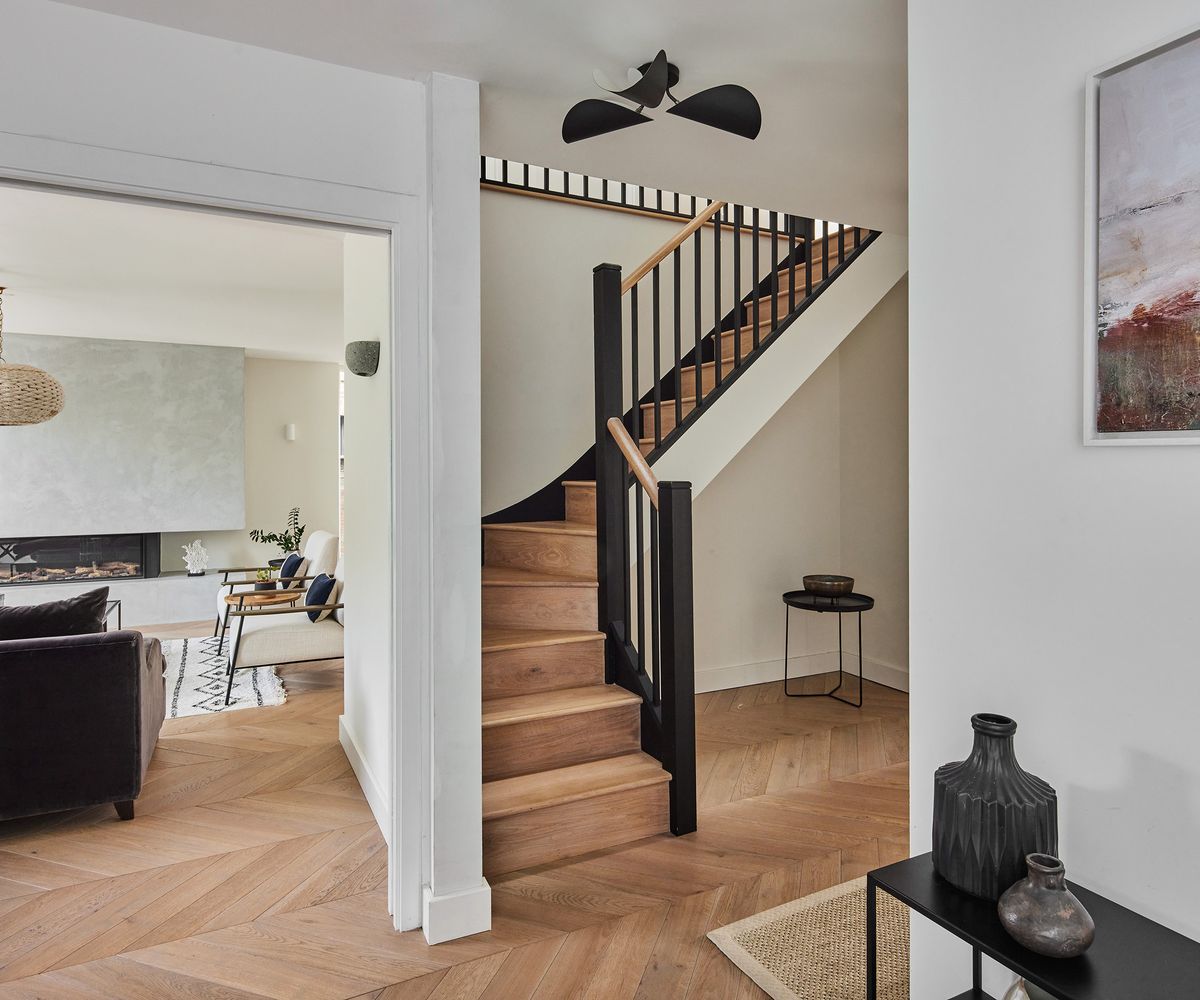 A hallway with white walls and engineered wood floor laid in a herringbone pattern. There is a doorway to the left with a view of a living room
