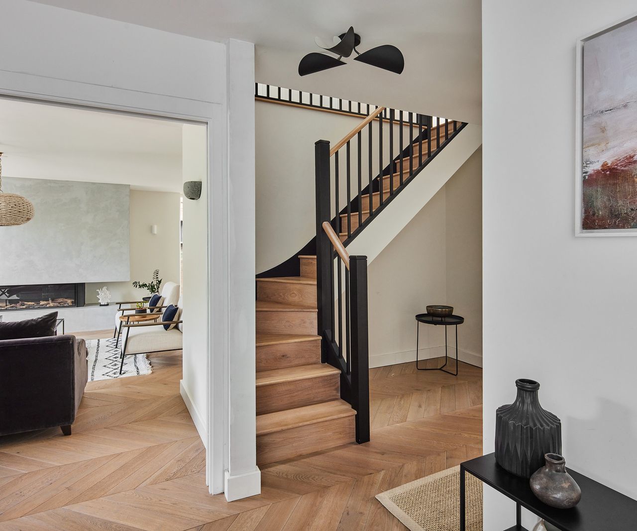 A hallway with white walls and engineered wood floor laid in a herringbone pattern. There is a doorway to the left with a view of a living room