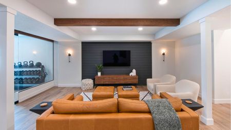 White basement with an entertainment center mounted on paneled dark gray wall, orange sofas and cream boucle armchairs, with ottoman orange footstools, and a gym visible to the left with dumb dumbbell rack and glass wall