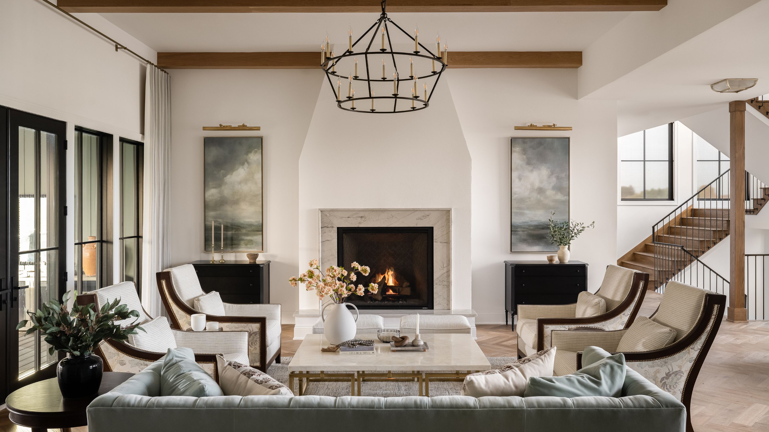 A symmetrical, bright living room with a central white fireplace, two matching black dressers with landscape art, four armchairs, and a soft blue tufted sofa in the foreground.