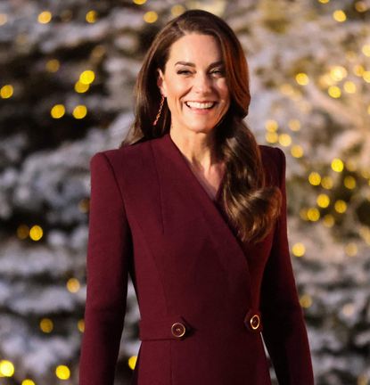 Kate Middleton smiling and wearing a burgundy coat in front of lit up Christmas trees