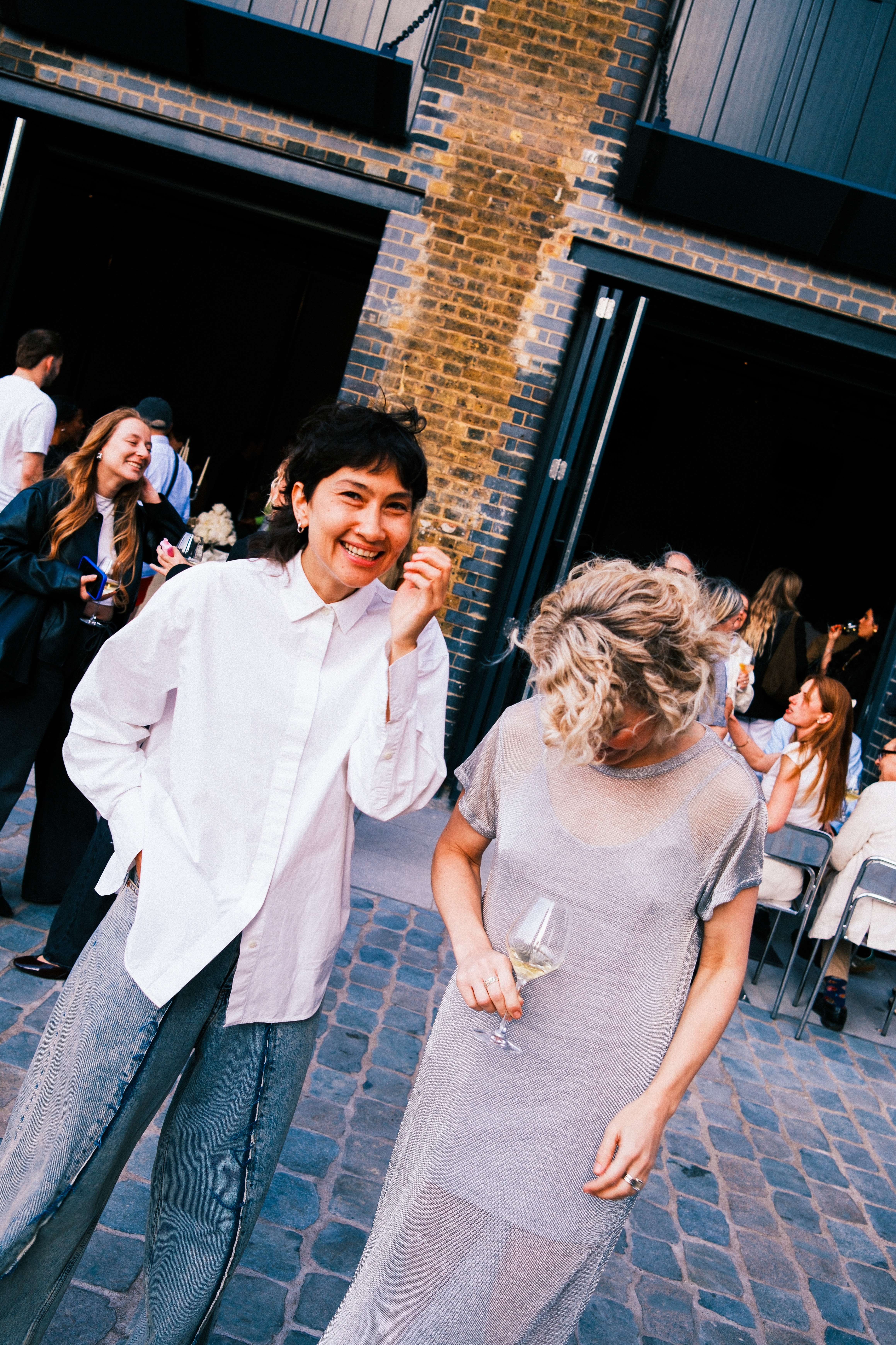 Two women dressed in smart clothes, one in a loose white shirt with detailed jeans, the other, in a see-through maxi dress in iridescent gray, stand jollingly in front of a brick-clad facade of a restaurant teeming with people.