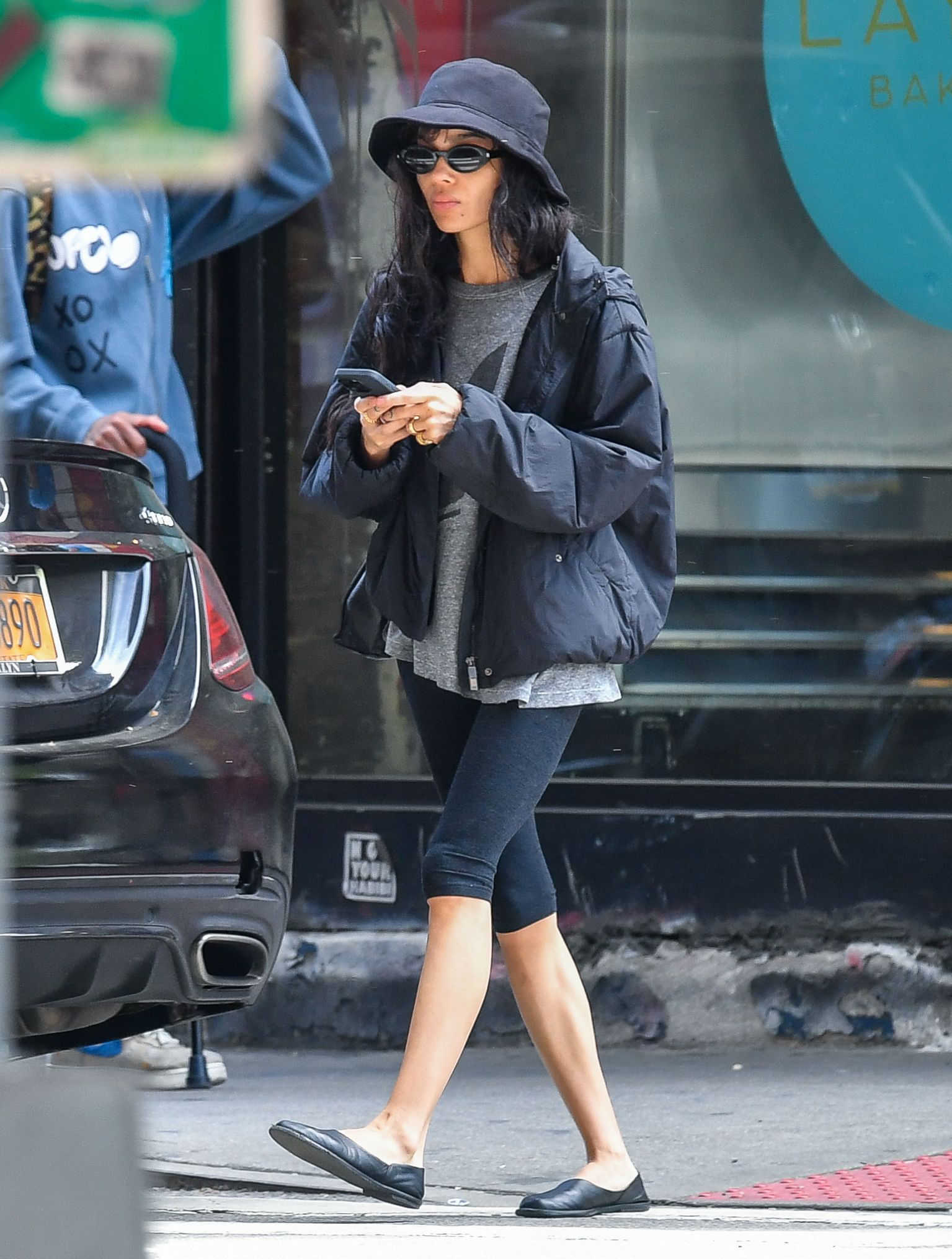 Zoe kravitz wears a black hat, gray t shirt, black jacket, black pedal pusher leggings, and black flats.