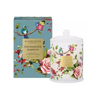 A white candle with an illustration of colorful birds, branches, and flower, with a teal box to the left of it with the same pattern and a gold label saying 'Glasshouse Fragrances Enchanted Garden'