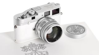 A silver and white Leica M camera with a matching silver lens rests on a white surface next to a round, silver lens cap engraved with a complex design and a papal coat of arms is visible beneath the camera.