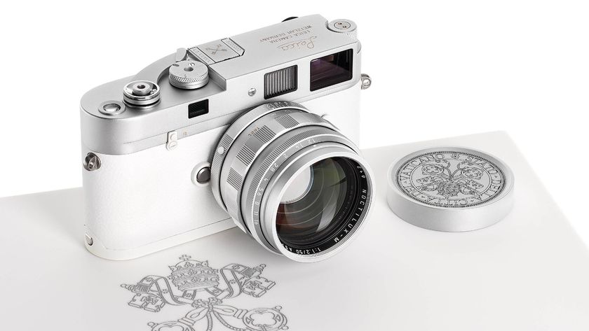 A silver and white Leica M camera with a matching silver lens rests on a white surface next to a round, silver lens cap engraved with a complex design and a papal coat of arms is visible beneath the camera.