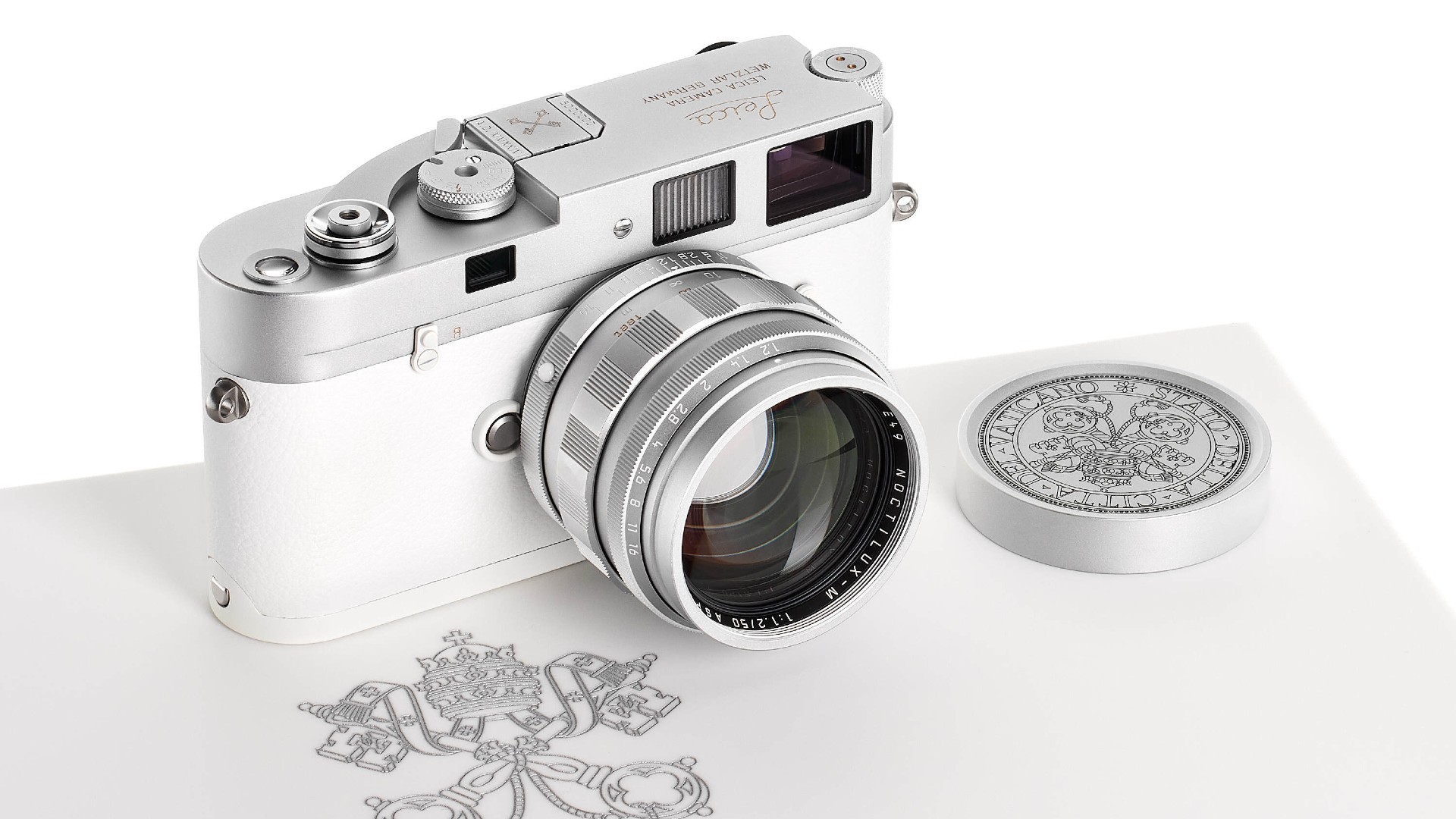 A silver and white Leica M camera with a matching silver lens rests on a white surface next to a round, silver lens cap engraved with a complex design and a papal coat of arms is visible beneath the camera.