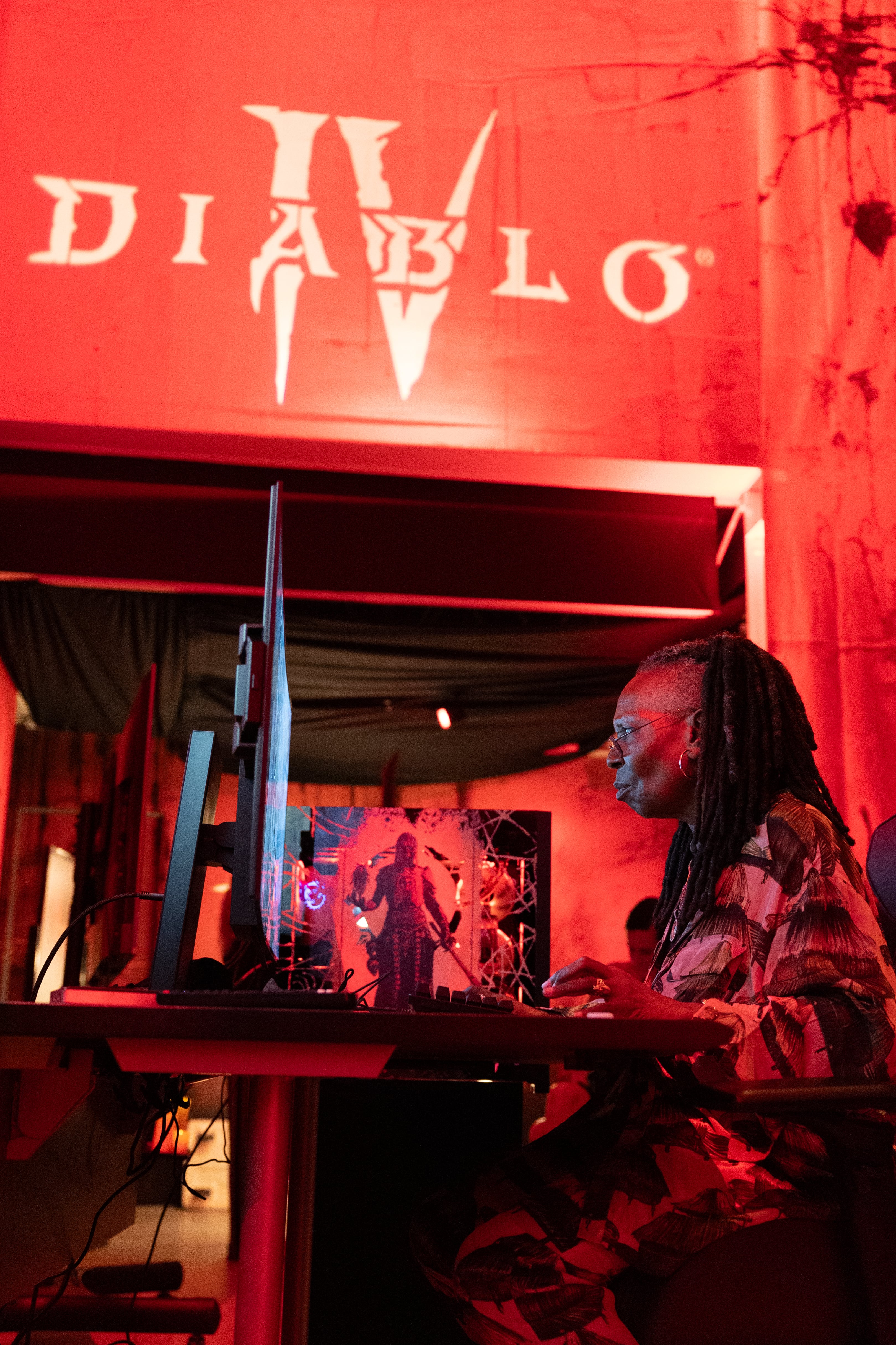 Whoopi Goldberg plays on a computer beneath a large Diablo 4 logo.