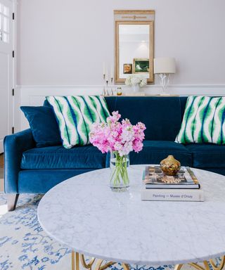 A white living room with a deep blue sofa, brightly colored green cushions, and a round white marble table with pink bouquet or flowers