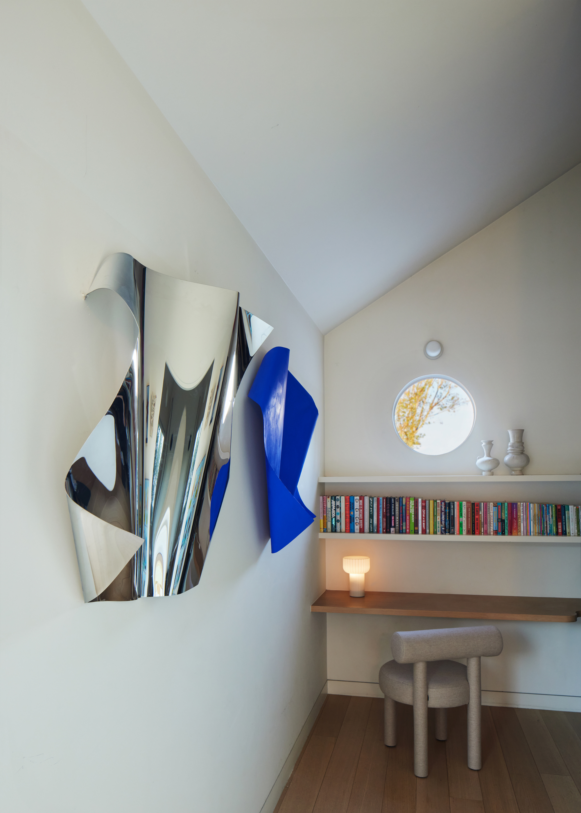 A white nook with a book shelf and a wood desk with a portable lamp and vases near a circular window and two textured chrome and cobalt blue wall hangings