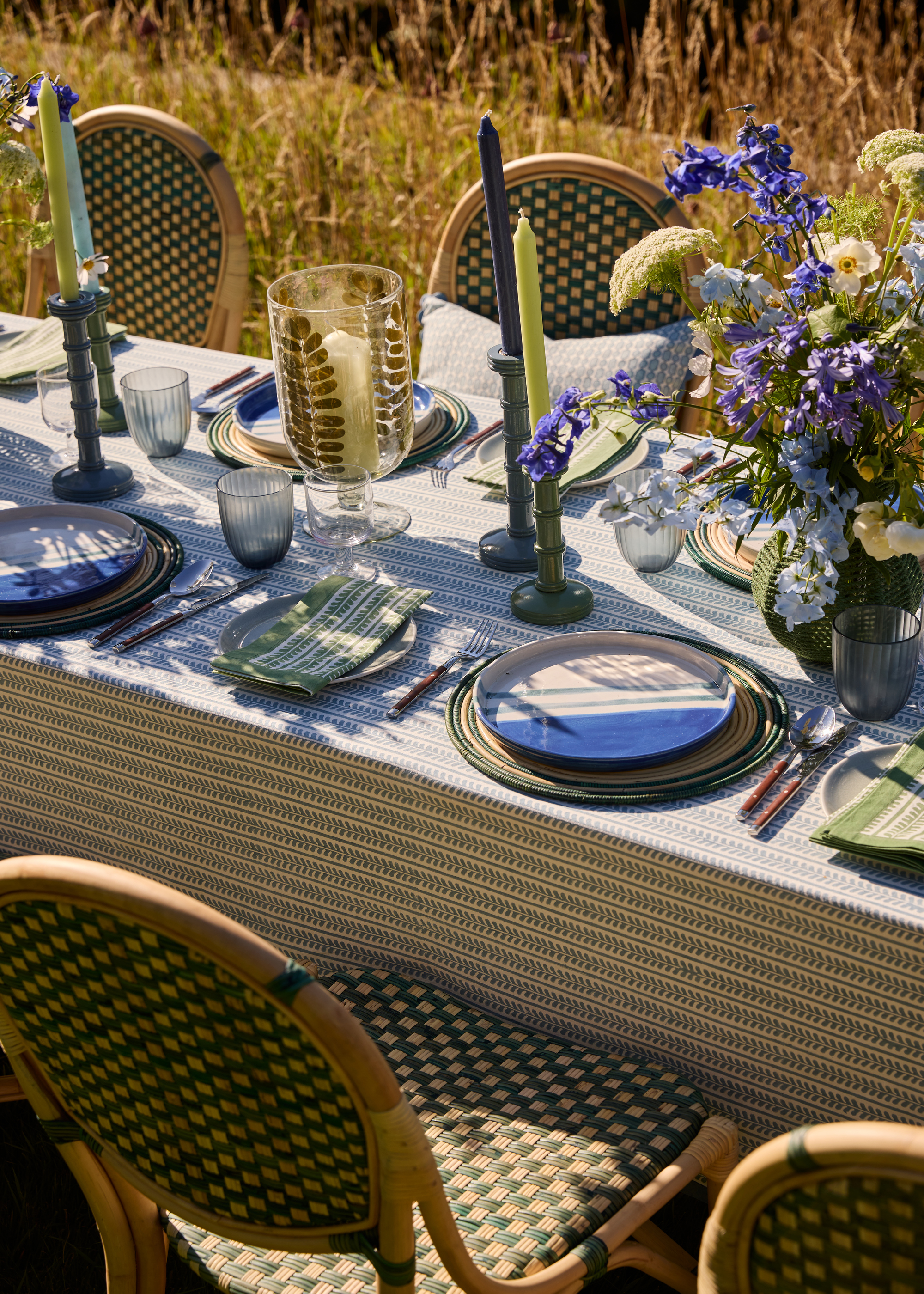 An outdoor dining table laid with green and blue themed decor, including candles, a vase of flowers, and striped napkins