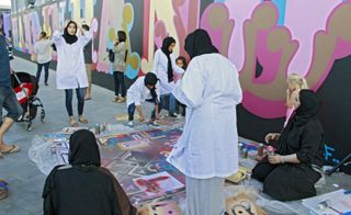 Women wearing hijabs are painting a mural that says 'Hand in hand'.