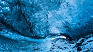 Inside Vatnajokull glacier with stunning blue-toned walls, shaped by glacial meltwater. The translucent ice forms intricate patterns, while snow and rocks cover the ground. Light filters through the entrance, creating a mesmerizing contrast between the icy textures and the cave’s depths.