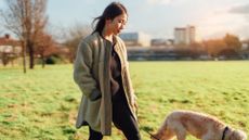 A woman walks her dog through a city park. The woman has black hair and wear a long coat. The dog is white and orange. The city skyline is visible in the background