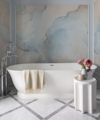 bathroom with a white freestanding bathtub, a small white side table with a candle and flowers and the walls painted with a blue and pink cloud mural