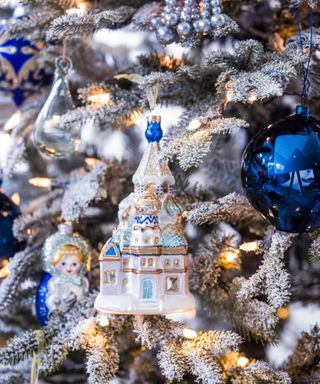 Close up of a Christmas tree, pine needles frosted with white artificial snow, blue and silver baubles, and ceramic ornaments, cherubs and Russian traditional building shapes