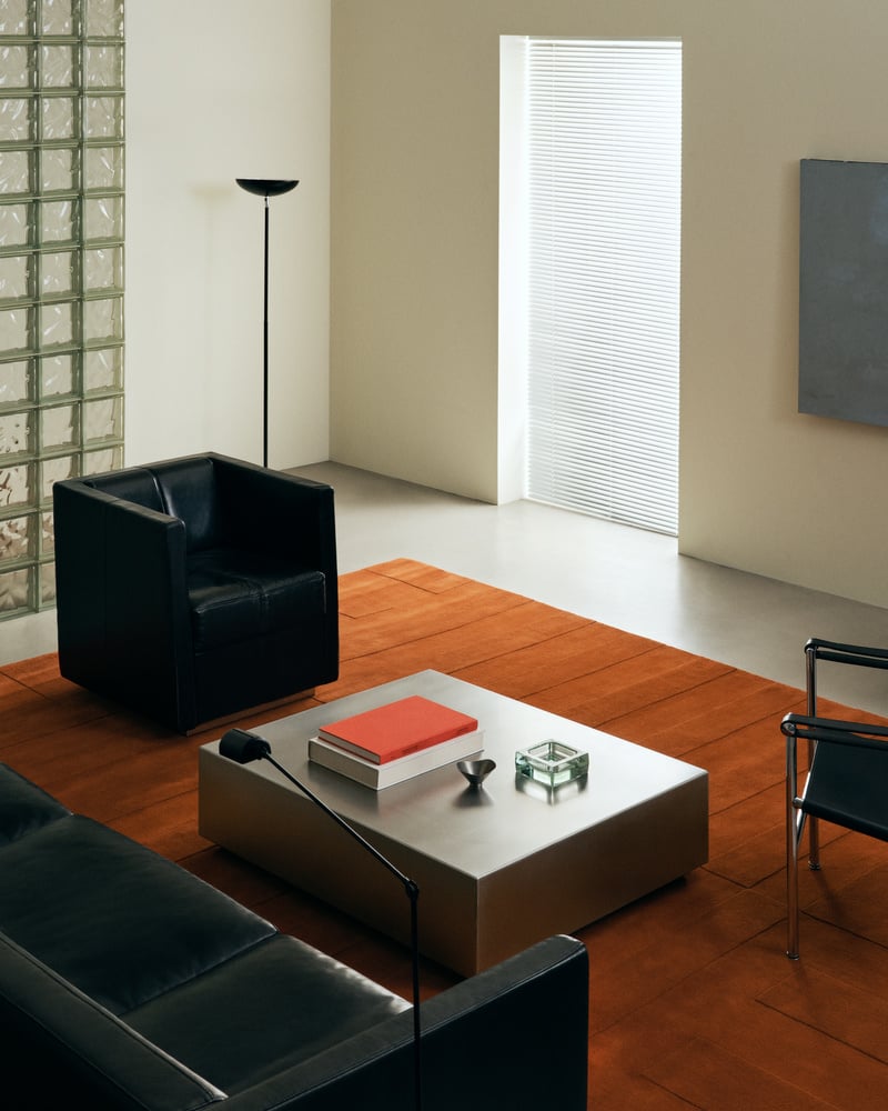 image of a modern living room with black leather seating and a rust orange rug. There is a chrome coffee table in the center of the room.