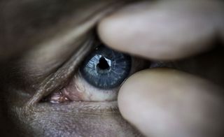 Close up of a blue eye, finger and thumb holding it open, reflection of the person taking the photograph in the pupil