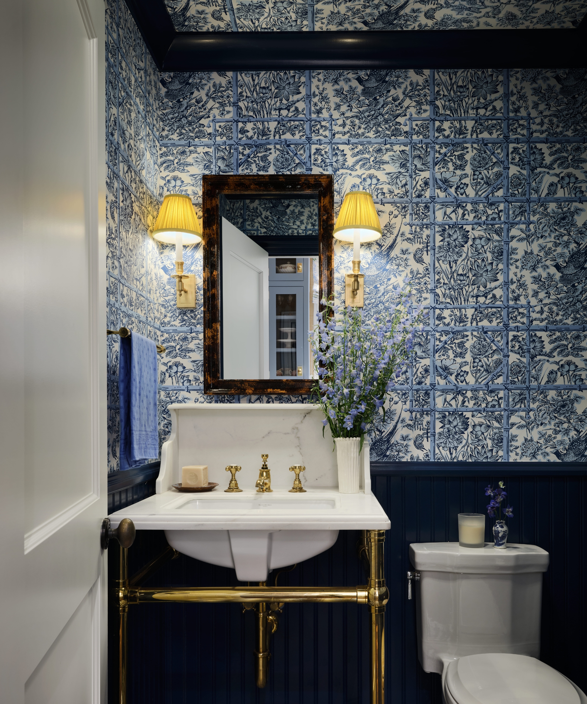 Blue-and-white powder room with botanical wallpaper, navy wainscoting, a marble basin and polished brass fittings.