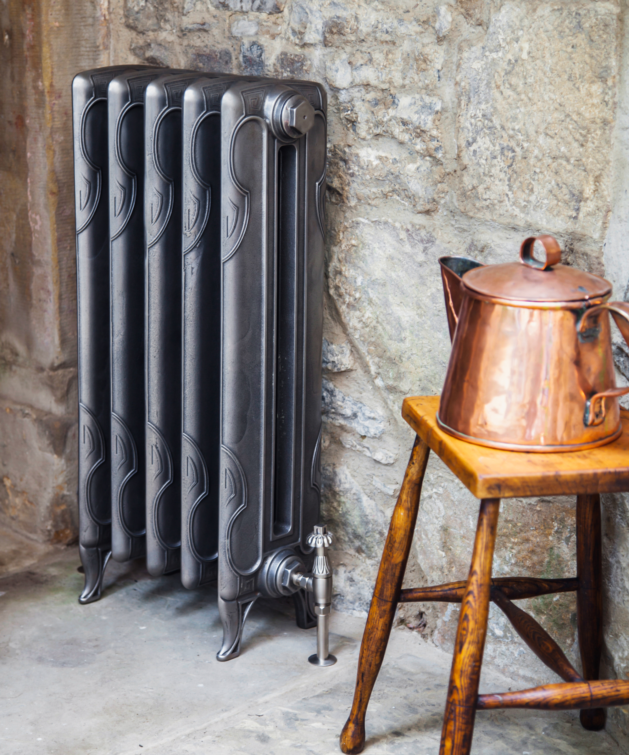 grey cast iron radiator against stone wall, with wooden stool and copper kettle stood next to it