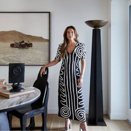 Image of a woman in a black and white dress standing in a modern room with a marble table and black chairs, a tall black floor lamp, and a desert photo on the wall.