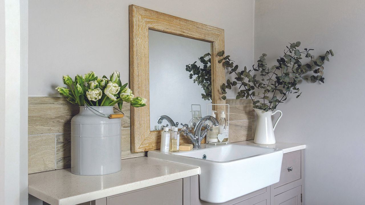 Bathroom detail with mirror over grey washstand with butler&#039;s sink, flowers in vintage enamel jug and milk bucket.