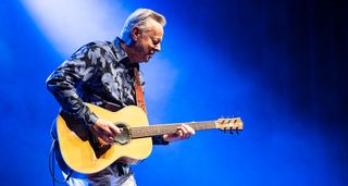 Against a blue backdrop, Tommy Emmanuel plays an orchestra-model sized acoustic guitar