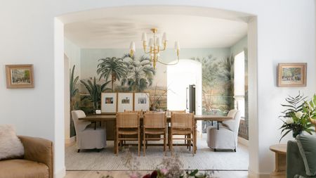 White interior archway revealing dining room with floral scene wallpaper and dining table with rattan chairs on white rug. Pictured in the foreground are an armchair, two framed pictures, and a side table with flowers.