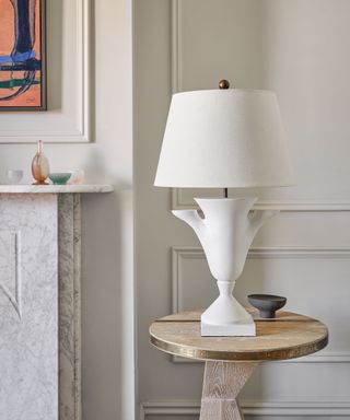 Close up of cream table lamp with sculptural, ceramic base, placed on top of wooden side table, artwork and fireplace in background