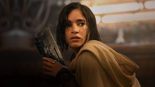 Still from the movie Rebel Moon (2023). Here we see a close up of Kora, a young woman with dark, chin length hair and wearing a beige cloak. In her hands she is grasping a large futuristic pistol.
